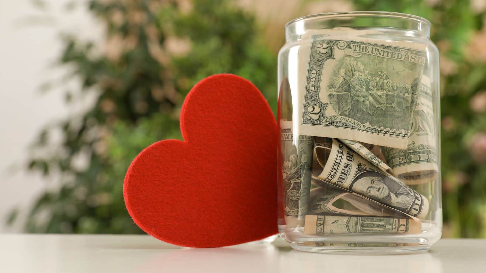 Image of a clear jar full of US dollars sitting next to a red felt heart